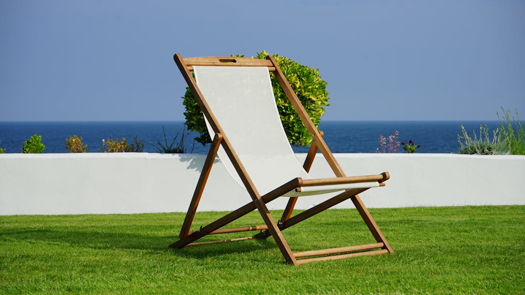 A wooden deck chair on lush green grass with a view of the blue ocean, perfect for a summer retreat.